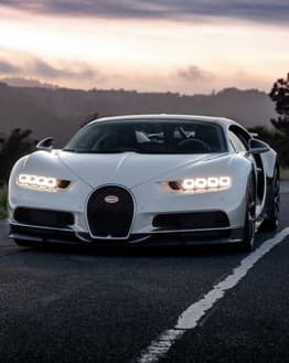Front view of a white Bugatti Chiron on a scenic road at dusk, featured on a watch face.