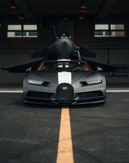 A matte black Bugatti Chiron with a white stripe, parked in front of a fighter jet in a hangar, featured on a watch face.