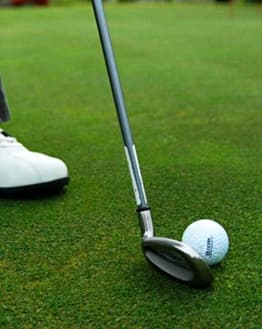 Close-up of a golf club and ball on a lush green with a player’s white shoe, displayed on a watch face.