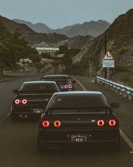 Photograph of three black JDM sports cars with iconic round taillights driving through scenic mountain roads at dusk, featured on a watch face.