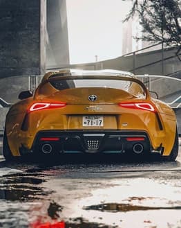 Photograph of a modified yellow JDM Supra sports car from the rear, showcasing its aggressive stance, glowing taillights, and urban reflections on wet pavement, featured on a watch face.