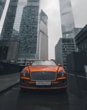 Front view of a bold orange Bentley Continental GT against a cityscape with skyscrapers, featured on a watch face