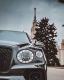 Close-up view of a Bentley Bentayga front headlight and grille with trees and a classic building in the background