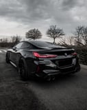 Rear view of a black BMW M8 under a cloudy sky, featured on a watch face