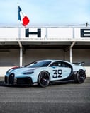 A Bugatti Chiron in light blue racing livery with the number 32, parked at a historic track with a French flag in the background, featured on a watch face.