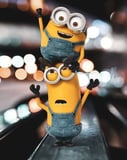 Two Minions dressed in blue overalls, stacked on top of each other with playful expressions, set against a blurred city light background, featured on a watch face.