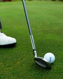 Close-up of a golf club and ball on a lush green with a player’s white shoe, displayed on a watch face.