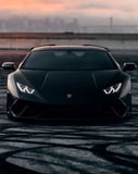 Matte black Lamborghini Huracán front view with a stunning sunset and urban skyline in the background, featured on a watch face.