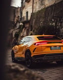 Rear view of a yellow Lamborghini Urus SUV parked in a rustic European-style city, showcasing luxury and performance, featured on a watch face.