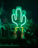 Glowing green neon cactus with a yellow pot, surrounded by potted plants in a dimly lit setting, featured on a watch face.