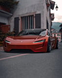 Tesla Roadster watch face displaying a bold orange Tesla Roadster parked on a quiet street surrounded by scenic buildings