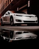 A Tesla Model S watch face featuring a white Model S in an urban alley with its reflection on a puddle, highlighting modern elegance.
