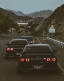 Photograph of three black JDM sports cars with iconic round taillights driving through scenic mountain roads at dusk, featured on a watch face.