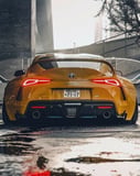 Photograph of a modified yellow JDM Supra sports car from the rear, showcasing its aggressive stance, glowing taillights, and urban reflections on wet pavement, featured on a watch face.