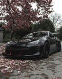 Photograph of a blacked-out JDM sports car with aggressive styling parked under cherry blossom trees with fallen petals, blending performance and Japanese aesthetics, featured on a watch face.