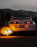 Photograph of a white JDM Supra sports car with glowing taillights and flames bursting from the exhaust, set against a dusk mountain backdrop, featured on a watch face.