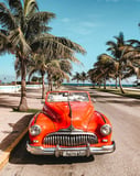 Photograph of a classic red convertible parked under palm trees on a sunny coastal road, blending vintage car culture with tropical vibes, featured on a watch face.