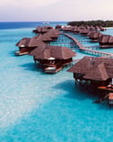 Aerial photograph of tropical overwater bungalows with thatched roofs, extending over crystal-clear turquoise waters in a paradise island setting, featured on a watch face.