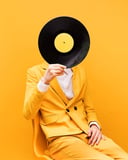 A person in a yellow suit sitting on a chair, holding a vinyl record as their face against a yellow background, designed as a stylish watch face